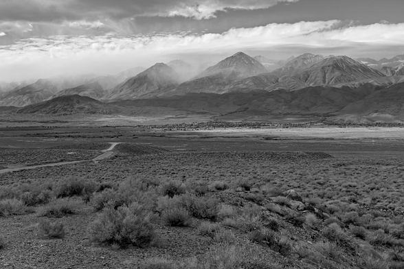 A black and white landscape photo of a tall rugged mountain range with numerous snow squalls covering some of the peaks and filling the valleys. In the middle ground is a small area of trees which mark the location of a small town. In the foreground is a vast plain of sagebrush. On the left side of the frame a road can be seen snaking down toward the distant town. The sky is mostly cloudy.