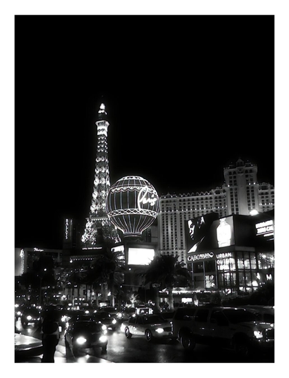 Black-and-white view of a bustling Las Vegas street at night, featuring illuminated Eiffel Tower replica and vibrant neon signs, evoking excitement.
