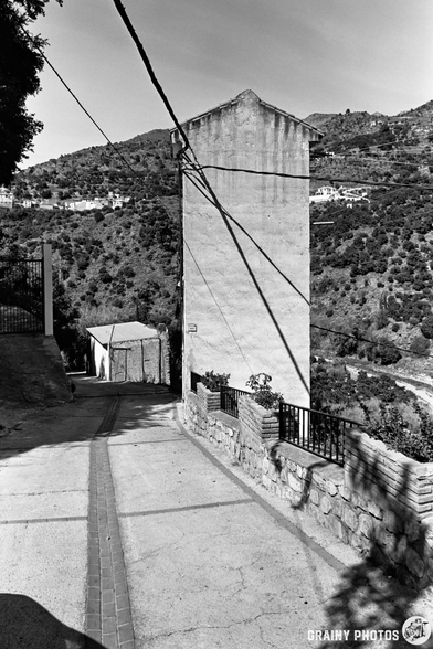 A narrow concrete road leads downhill, bordered by a stone wall and metal fence, towards a tall, narrow substation near Guájar-Fondón, set against a hilly landscape with scattered houses. The image is in black and white.