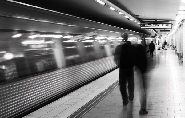 An black and white image taken on a subway platform as a train zips past, with some people walking. All moving elements have motion blur.