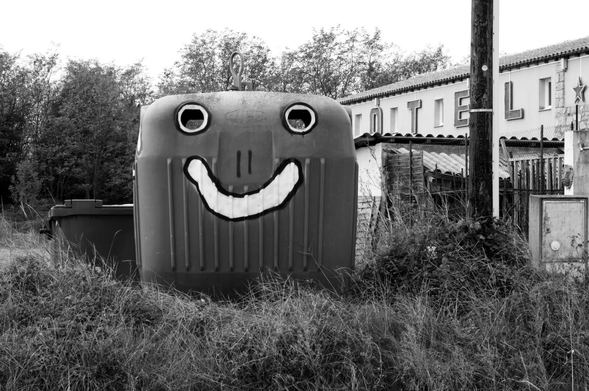 A street artist transformed a glass recycling bin into a face by painting white circles around the holes to create eyes and drawing a big smile underneath. (Version françæ) Æn artiste dal vie a taggé æn poubelle de recyclage pour læ verre en entourant les trous de blank pour faire des yeux et en dessinant æn granx sourire en dessous.