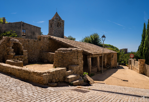 Color photo of an old Catalonian village under blue skies, with a small raised terrace next to a cobblestone street containing stone well and water trough structures and beyond it is a low tile-roofed structure fronting on an unpaved street. In the background is a square church tower. 