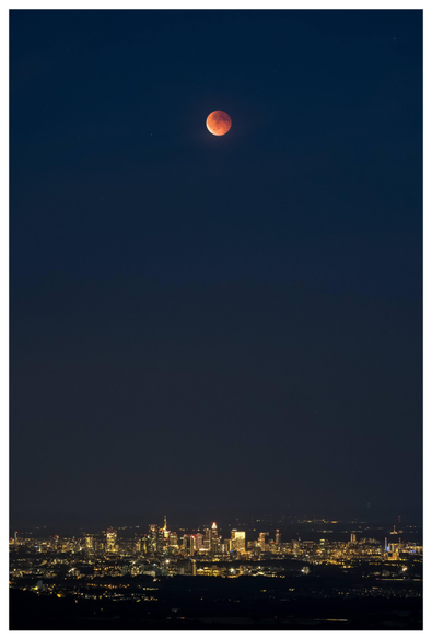 A vertically taken photo showing the blood moon high above over Frankfurt's illuminated skyline.