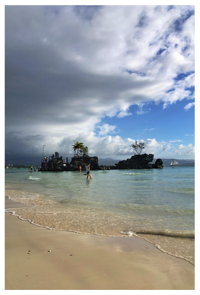 Photo of a tropical island beach, looking out to sea. A stretch of fine sand in the foreground leads to shallow water, with people wading calf-deep in the waves. In the middle distance offshore is a long, low rock formation topped by some trees. At its leftmost section is a small grotto (with a statue of the Virgin Mary inside, unseen), with people climbing up a short flight of steps from the water to reach it. A sailboat glides along the water further in the distance. Mountains lie on the horizon, under masses of low clouds sweeping across the blue sky.