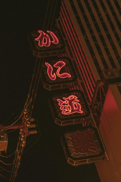 A vertical neon sign with glowing red Japanese kanji characters illuminates the night sky on a city street, attached to the side of a tall building with power lines visible nearby. Taken in Osaka, Japan.