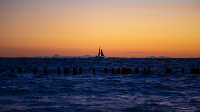 Farbfoto im Querformat. Sonnenuntergang über der Ostsee, im Vordergrund Wasser und eine Buhnenreihe, am Horizont ein kleines Segelboot vor orangefarbenem und ross Himmel und ein paar Wolken.