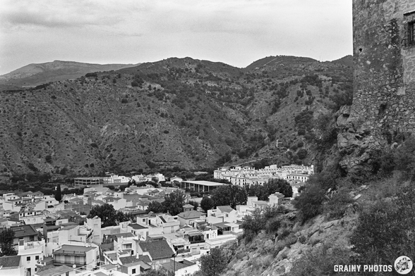 Black and white photo of Vélez de Benaudalla viewed from the castle, showing tightly packed hillside buildings, mountains in the background, and part of Castillo de Vélez de Benaudalla is visible on the right.