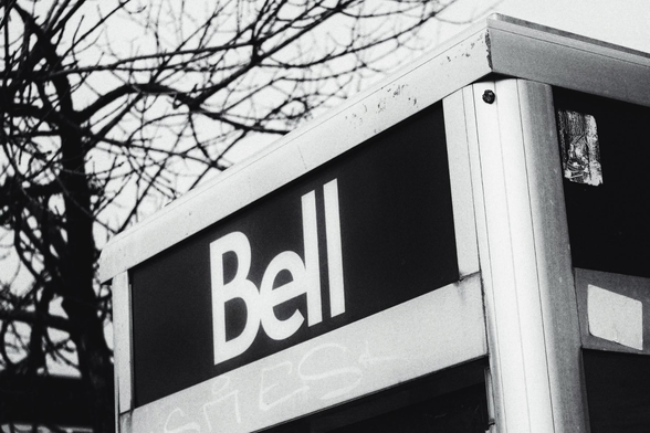 The top edge of a phone booth with the word "Bell" written on it is seen in this black and white image. A large leafless tree is seen in the background.