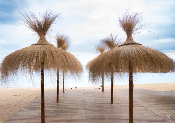 Color photo looking through a double row of beach umbrellas made of raffia-like leaves towards the beach and the Mediterranean Sea. 