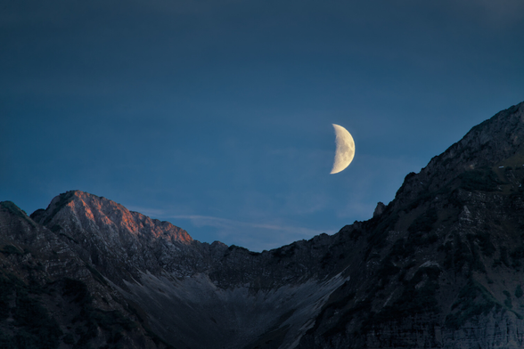 A dark mountain saddle where the moon goes up above. The sky is turning dark blue, the top of one mountain to the left is still getting lit by the sun, giving some Alpenglow to the scene