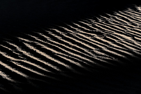 A black and white landscape photo of ripples in the sand dune. The editing has made the photo very high contrast with black borders and a beam of light shinning in from the lower left to the upper right. The ripples are very prominent with nearly black backsides and very bright front sides where the low angle sunlight was hitting them.