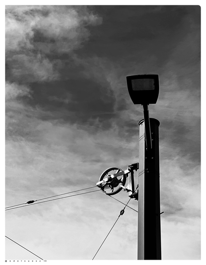 Schwarzweißfotografie: hoher dunkler Stahlmast mit zusätzlicher Straßenbeleuchtung, oberhalb ein Radspanner mit Verzahnung außen, diese werden gebraucht um die Oberleitungen gespannt zu halten, grauer Himmel im hellen Wolken im Hintergrund