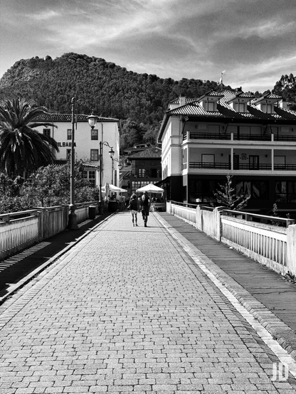 Fotografía en blanco y negro de un pintoresco paisaje urbano. En primer plano, un puente peatonal adoquinado se extiende hacia el fondo, flanqueado por barandillas blancas de madera. Dos personas caminan de espaldas, alejándose del espectador por el centro del puente.

A los lados, se aprecian edificios tradicionales: a la izquierda, uno blanco con el letrero "BILBAINA" y una palmera; a la derecha, otro de varios pisos con un distintivo tejado de tejas, buhardillas y balcones con barandillas oscuras. Varias farolas ornamentadas se distribuyen a lo largo del puente y las calles adyacentes, donde también se ven sombrillas blancas de establecimientos.

Al fondo, una imponente montaña densamente boscosa domina el paisaje, bajo un cielo parcialmente nublado.