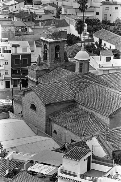 Black and white photo of Vélez de Benaudalla, highlighting the central stone Iglesia Nuestra Señora del Rosario with its bell tower and dome, surrounded by tiled-roofed houses and leafy trees in the background.