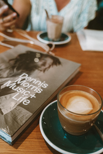 Photo of a café table with a coffeetable book showing a photo of Bob Marley. The book is the exhibition catalogue of Dennis Morris Music + Life. In the foreground is a cup of coffee. In the background a woman with a café latte and a phone in hand. 