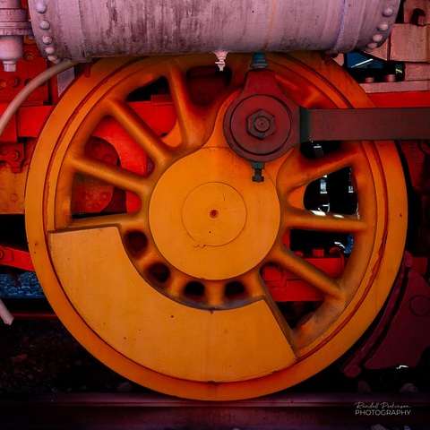 A steam locomative drive wheel painted bright orange along with other portions of the locomotive painted pink, burgundy, and another shade of orange to highlight different parts of a steam locomotive for guests at a transportation museum.