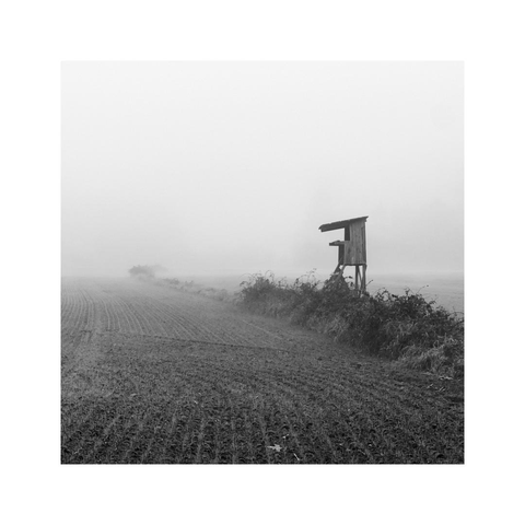 the landscape is disappearing in the distance in a wall of dense, white fog. at midrange there are the outlines of a black deerstand on a wide field still clearly visible. some small bushes seem to guide your way to that so marked place in the distance