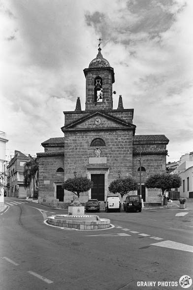 A black and white photo of Iglesia Nuestra Señora del Rosario in Vélez de Benaudalla shows its stone bell tower, parked cars in front, trees on either side, and a cloudy sky as the street curves round a small roundabout in the foreground.