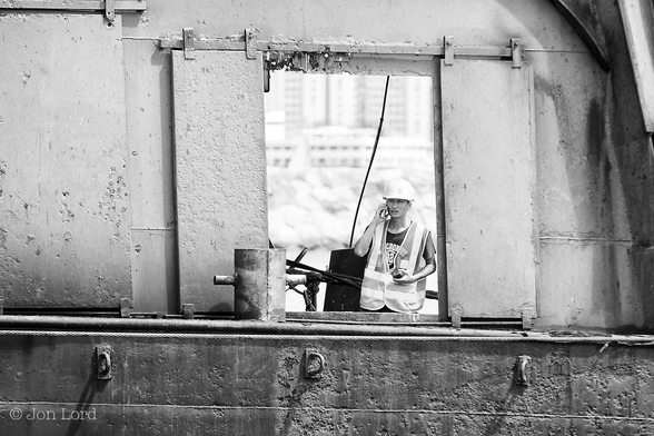 This is a black and white street photo in landscape format of a young Chinese engineer on a maritime construction site. Causeway Bay, Hong Kong (2014).
Stretching along the lower quarter of the image is the black and pitted steel hull of a derrick barge (a large square shaped barge with a deep hull and a large crane at one end on top of a two floor superstructure), with a thick welding seam running horizontally across with three large steel mooring or attachment eyes above. On the narrow deck is a large bollard with a taut steel hawser stretched across the frame from left to right. Above the deck is a medium grey superstructure, also pitted and with multiple welding seams running horizontally. In the centre of the image are two open steel shutters of about two metres tall by half a metre wide. Behind this opening is a young Chinese engineer wearing a high-vis tabard, a t-shirt beneath and a white safety helmet on his head. He is holding a mobile phone in his right hand to his right ear and holding a small two-way radio in his left hand. Our engineer is looking out of the opening and slightly to the left of the camera. The background is heavily blurred out of focus with a breakwater in view and high-rise buildings in the distant behind. 
This was the scene of construction of the Central- Wan Chai Bypass, in this part, a tunnel under the Causeway Bay Typhoon Shelter. 