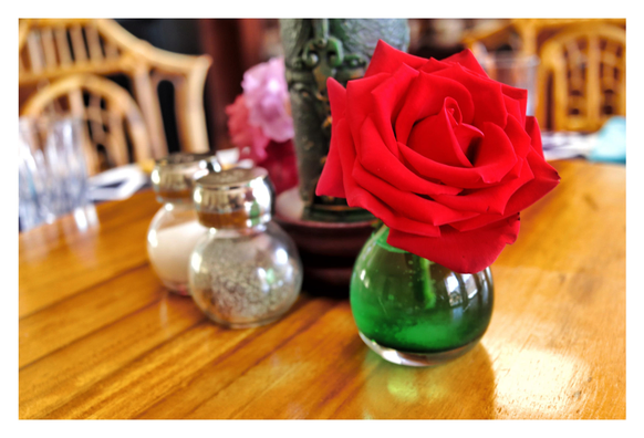 Photo of a perfect red rose with velvety petals sitting in a small, round, green vase, on a Lazy Susan with a glossy sheen over its wood grain. Behind and to the left of the rose are salt and pepper shakers and a large green decorative candle with a molded surface pattern. Further back are drinking glasses and pink and purple blossoms in vases, and at the edge of the dining table are rattan or bamboo chairs. Everything is slightly out of focus except for the rose.