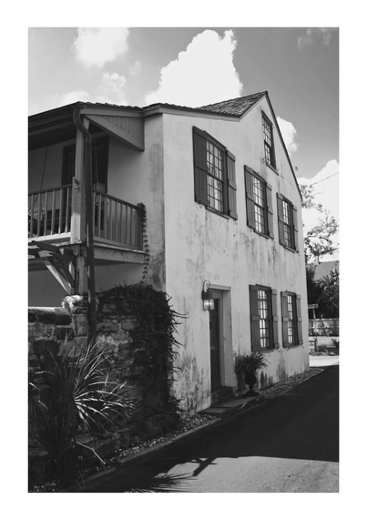 A monochrome image of an old, two-story house with shuttered windows and a small balcony. It's located on a quiet street, evoking a nostalgic feel.