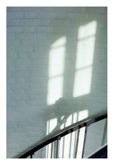Sunlight casts window shadows on a white brick wall beside a curved metal staircase, creating a calm and serene atmosphere.