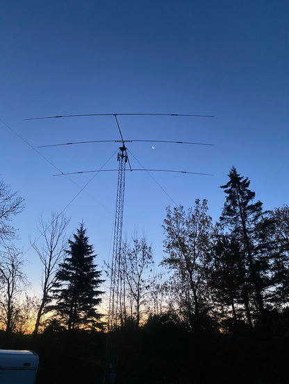 Bright crescent moon between the elements of a large backlit three-element ham radio antenna on a metal tower. Dark shapes of bare and coniferous trees along the horizon, and the light fades from orange below to dark blue above.