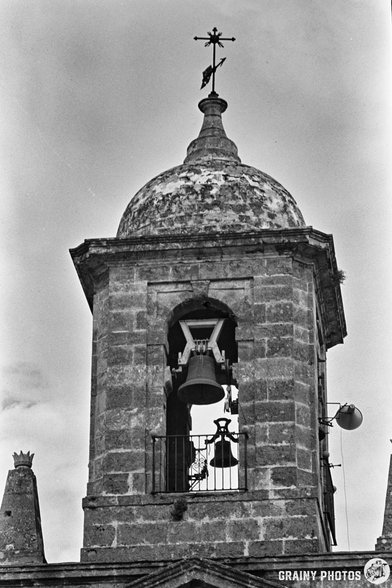 Black and white photo of the Iglesia Nuestra Señora del Rosario’s stone bell tower in Vélez de Benaudalla, topped with a metal cross and visible bells. Weathered surfaces stand out against a cloudy sky.