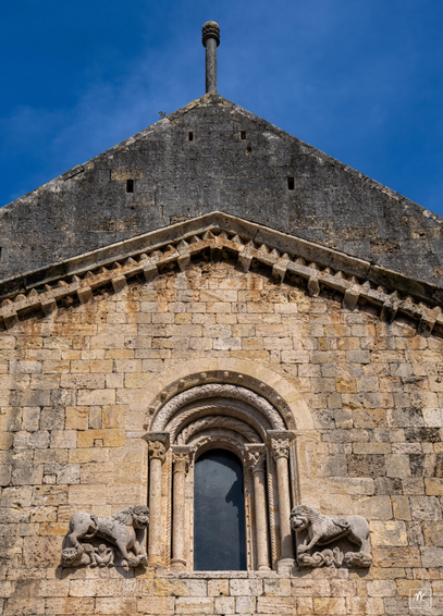Color photo of the upper part of the gable end of an old stone choice building with a single central inset arched window flanked by fanciful medieval lion sculptures on either side. 