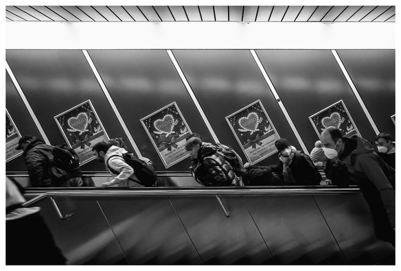 Menschen auf der Rolltreppe. So fotografiert, dass die Treppe parallel zu den Bildrändern ist und die Menschen daher schräg zu stehen scheinen.