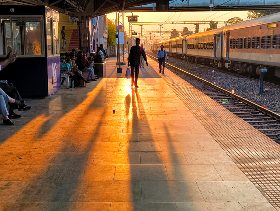 Long shadows stretch across a train station platform as the setting sun casts a golden glow. A train is visible on the tracks to the right, and people are waiting on the platform, some seated, others walking. The warm light highlights the figures and the texture of the platform.