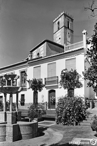 A black and white photo of a traditional Spanish-style building in Vélez de Benaudalla showcases a tower, balconies, and a tiled roof. A stone fountain and lush plants fill the foreground. A church bell tower is visible in the background, set against clear skies above.