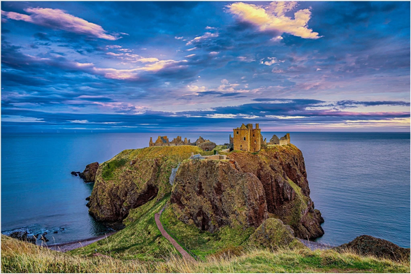 A coastal landscape featuring the ruins of a castle atop a grassy cliff, overlooking a calm sea. The sky is dramatic with colorful clouds, suggesting sunset or sunrise. A pathway leads down towards the castle.