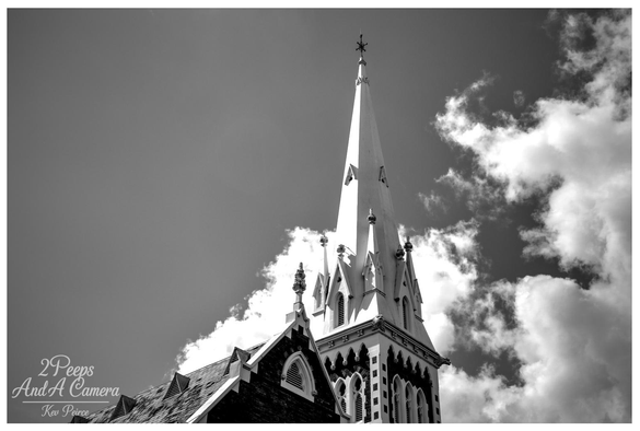 A high contrast black and white photograph, signed by Kev Peirce, focusing on the tall, white spire of a historic church in Launceston, Tasmania.

The spire dominates the frame, rising sharply against a dramatic sky with bright, billowing clouds.

The dark roof and upper structure of the brick church are visible below the spire, framed from a low angle.