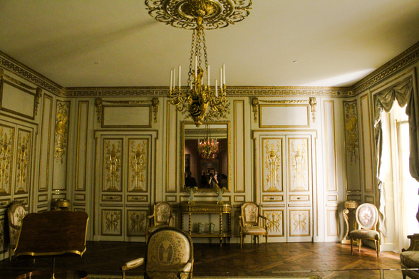 A wide shot of various chairs and neoclassical furniture in a sunlight room. The wall panels and doors are cream with gold borders.