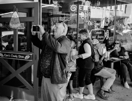 A black and white photo of a guy taking a photo using his cell phone.  There are people entering a candy store behind him and a couple sitting at a table right outside the store.