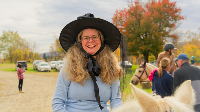 Woman with lots of blond hair wearing a blue blouse and a black witch hat who is riding a horse whose ears you can barely see at the bottom right.