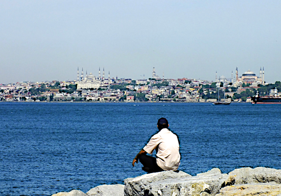 Foto van gehurkte man die over het blauwe water van de Bosporus naar het Europese deel van Istanbul kijkt