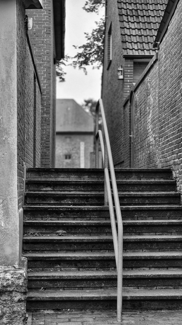 Auf dem Schwarzweißfoto ist ein Treppenaufgang mit einem Mittelgeländer aus Metallrohr zwischen zwei Häuserwänden zu sehen. Die Häuser sind aus Ziegelsteinen gebaut. Am Ende des Durchgangs sind ein weiteres Haus sowie Teile von Baumkronen zu sehen. Es ist eine triste, öde Szene, die im Alltag unterginge.