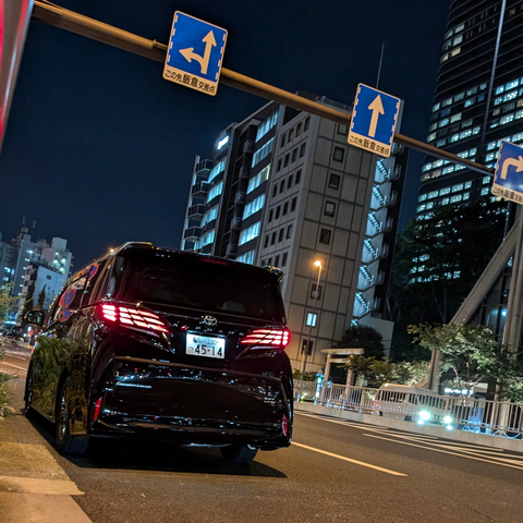 At night, a black toyota van sits at the side of the street with its rear lights on. Above it there's two street signs and in the background multiple mid sized high rises with its window lights on.
