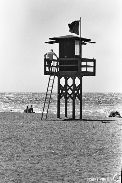 A lifeguard climbs a wooden lookout ower on Salobreña beach, with a flag flying above. The sea is visible in the background and a few people sit near the shore under the bright sunlight.