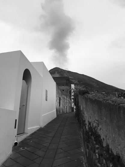 Une rue pavée sur les pentes du Stromboli est bordée sur sa droite d’un mur gris; à gauche, des mur d’habitation dont le premier est peint en blanc; en arrière plan, le cône du Stromboli crache une fumée noire