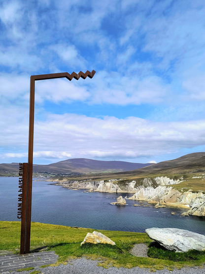 Hellblauer locker bewölkter Himmel und ein Blick auf weiße zerklüftete Klippen und sanfte graugrüne Berge im Hintergrund.
Das Meer zeigt sich in unterschiedlichen Blautönen. Im Vordergrund ist saftiges frisches Gras, zwei Felsbrocken in der Farbe der Klippen und auf der linken Seite ein Metall Pfahl des "Wild Atlantik Ways" mit dem gälischen Namen des Ortes, hochkant geschrieben. Im Himmel sieht man den Pfahl wie er nach rechts abknickt und dann zackig endet. Diese Zacken sind das Symbol des "Wild Atlantik Ways" und findet man an allen Wegpunkten.