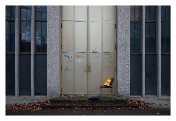 Farbfoto. Die hohe, zweiflügelige Portaltür einer Kirche geht oben aus dem Bild heraus. Sie ist aus Metall, mit Graffiti bespüht. Zwei flache Stufen führen zu ihr. Auf der oberen steht ein Veranstaltungststuhl mit Rohrgestell und braunen Polstern. Auf dem Stuhl steht eine große, gelbe Warnleuchte. Vor dem Stuhl liegt ein blauer Sitzkeil auf dem Boden. Auf und neben den Stufen ist feuchtes Laub.

Colour photograph. The tall, double-winged portal door of a church extends beyond the top of the picture. It is made of metal and covered in graffiti. Two shallow steps lead up to it. On the top step stands an event chair with a tubular frame and brown upholstery. A large yellow warning light stands on the chair. In front of the chair, a blue seat wedge lies on the floor. There are damp leaves on and next to the steps.