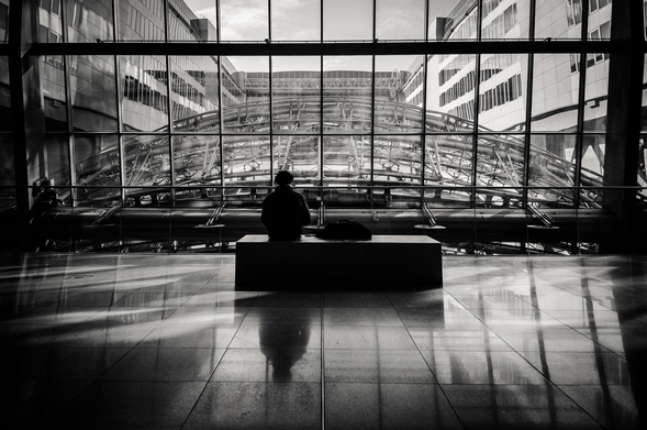 The image is a black and white photograph depicting a modern architectural interior. The primary focus is a person sitting on a dark rectangular bench, facing away from the camera and towards a large window.

The interior space appears to be a large hall, possibly a terminal or a public space within a modern building. The dominant feature is a massive glass window that spans the width of the frame. Through the window, a complex network of metal pipes and supports is visible, suggesting an industrial or infrastructural element beyond. The architecture visible through the window includes more contemporary buildings with glass facades and clean lines.