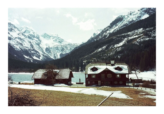 Snowy mountain landscape with a serene lake and two rustic wooden cabins in the foreground. The sky is cloudy, creating a tranquil, wintery atmosphere.