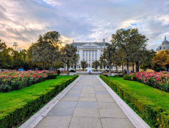 A symmetrical view down a stone walkway in a park in Zagreb. The path is flanked by green hedges and beds of vibrant pink roses, leading towards a fountain and a grand, white neoclassical building under a cloudy sky.
