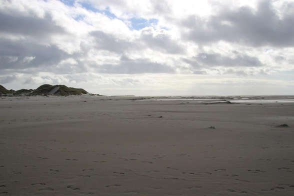 Blick über einen weiten Sandstrand, links erhebt sich eine Düne, rechts das Meer, darüber ziehen Wolken
