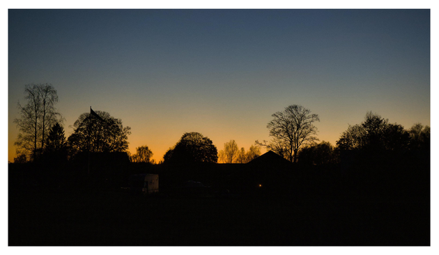 En liggande bild på silhuetten mot himmelen när solen gått ner och en orange rand fortfarande syns längst ner. Träd, några hus, en flaggstång och buskar skapar silhuetten. Ett fordon kan anas i skuggorna, några små ljus syns.