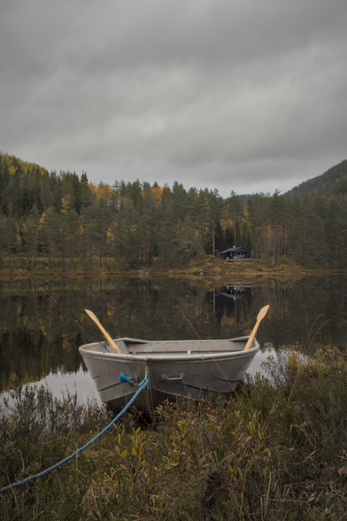 A small boat is in the foreground, with oars sticking out on each side. Behind it, on the other side of a small lake, is a forest and a small cabin. Everything has autumn colors. The sky is overcast. The lake is completely still, like a mirror. 